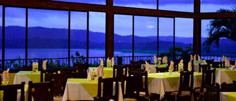 A restaurant with tables set for dining, large windows offering a view of the sea and mountains, and a tropical palm tree visible outside.