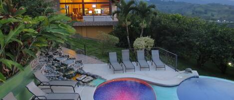 Un complejo turístico con piscina, sillas de descanso y un edificio con amplios ventanales con vistas a un paisaje montañoso.