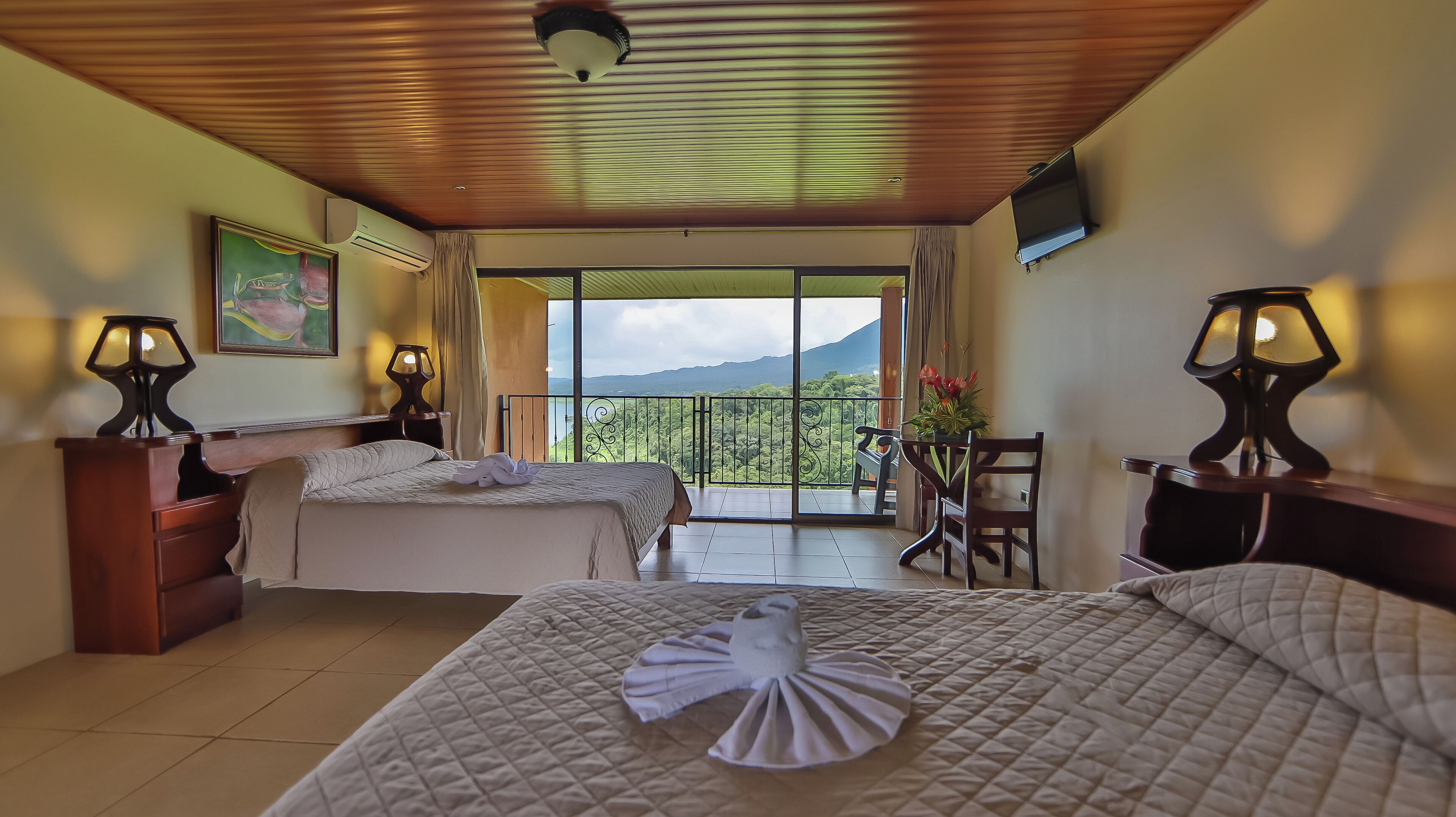 A hotel room with two beds, a balcony with a view, a dining area, and a TV mounted on the wall.