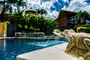 Une piscine agrémentée d’une sculpture en pierre, entourée d’une végétation luxuriante et bordée d’un bâtiment dont le toit est recouvert de tuiles.