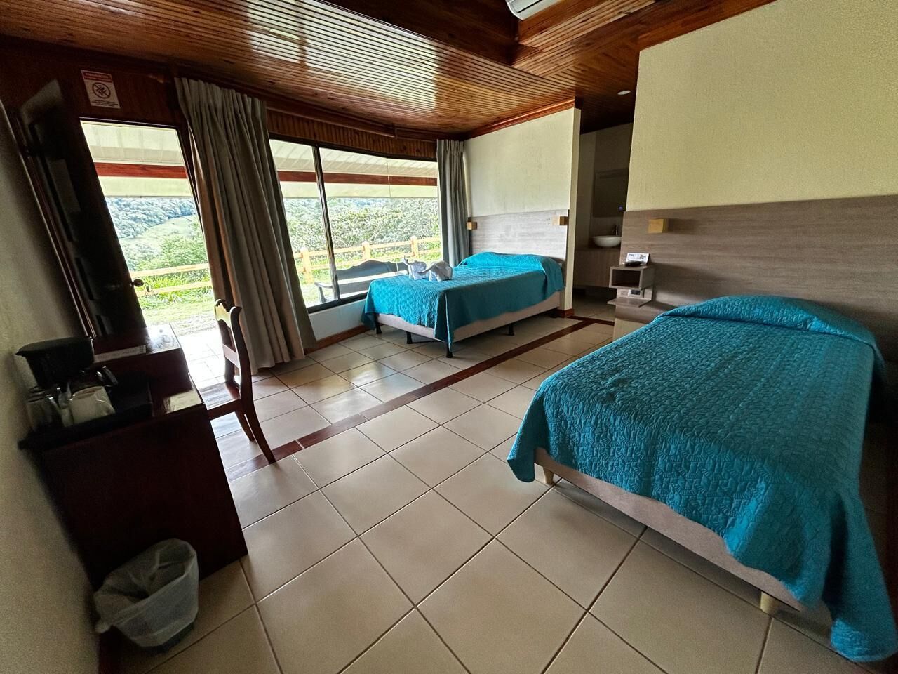 Quarto de hotel com duas camas, uma escrivaninha, uma cadeira e vista para o exterior.