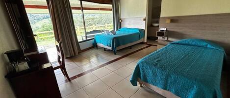 Quarto de hotel com duas camas, uma escrivaninha, uma cadeira e vista para o exterior.