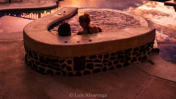 A hot tub with a stone base, surrounded by a pool, and overlooking a scenic view of a sunset over a body of water and hills. - Hotel Linda Vista (La Fortuna)
