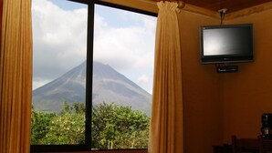 A hotel room with a wooden ceiling, a large window with curtains, a flat-screen TV, and a bed with towels and flowers. - Hotel Linda Vista (La Fortuna)