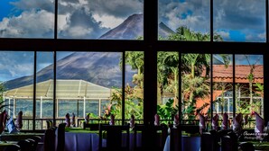 A restaurant with a view of mountains and a greenhouse. Tables are set with white tablecloths and purple napkins. - Hotel Linda Vista (La Fortuna)