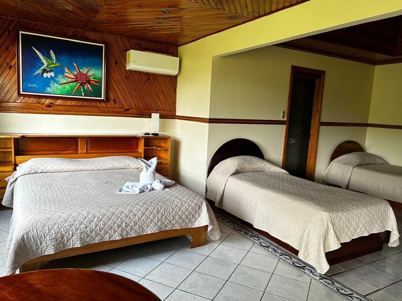 Habitación de hotel con techo de madera, un cuadro de colibrí y flor, dos camas con ropa de cama color crema, y un armario de madera.