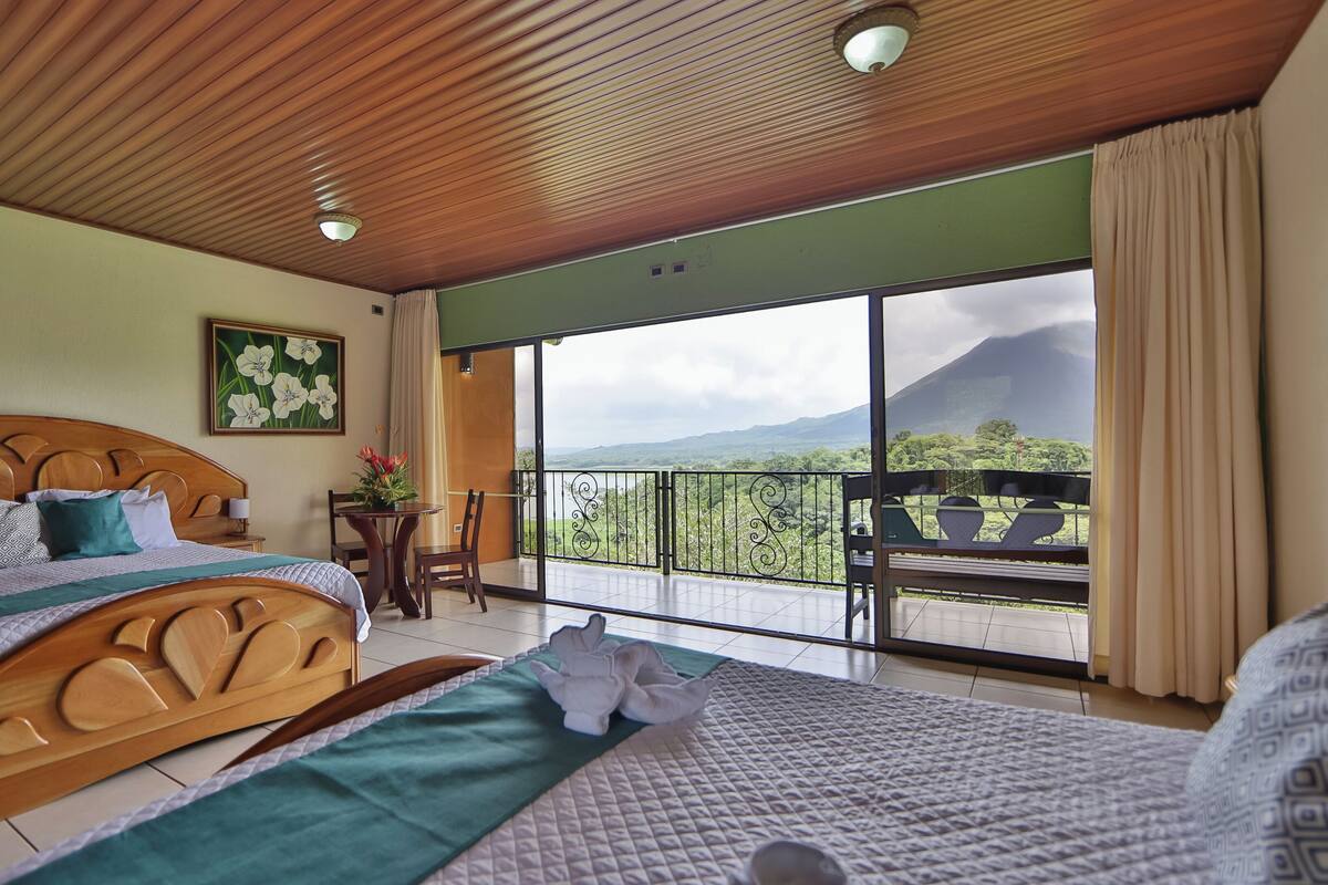 Een hotelkamer met een houten plafond, een groot bed met houten hoofdeinde, een eettafel met stoelen en een balkon met uitzicht op een berg.