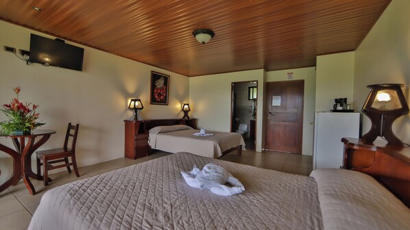 A hotel room with two beds, a wooden ceiling, a TV mounted on the wall, a desk with a lamp, and a door leading to another room.