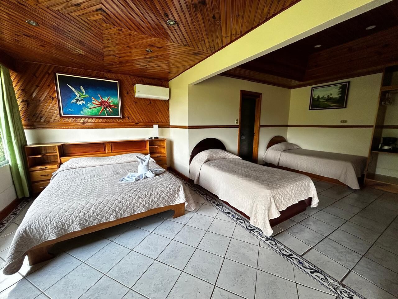 A bedroom with a large bed, two smaller beds, wooden ceiling, tiled floor, and artwork on the walls.