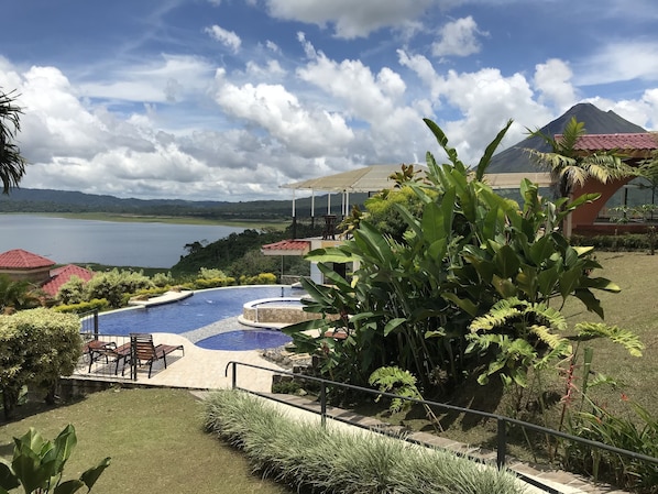 Un espace piscine offrant une vue sur un lac et des montagnes, entouré de plantes tropicales et bordé d’un bâtiment au toit rouge.
