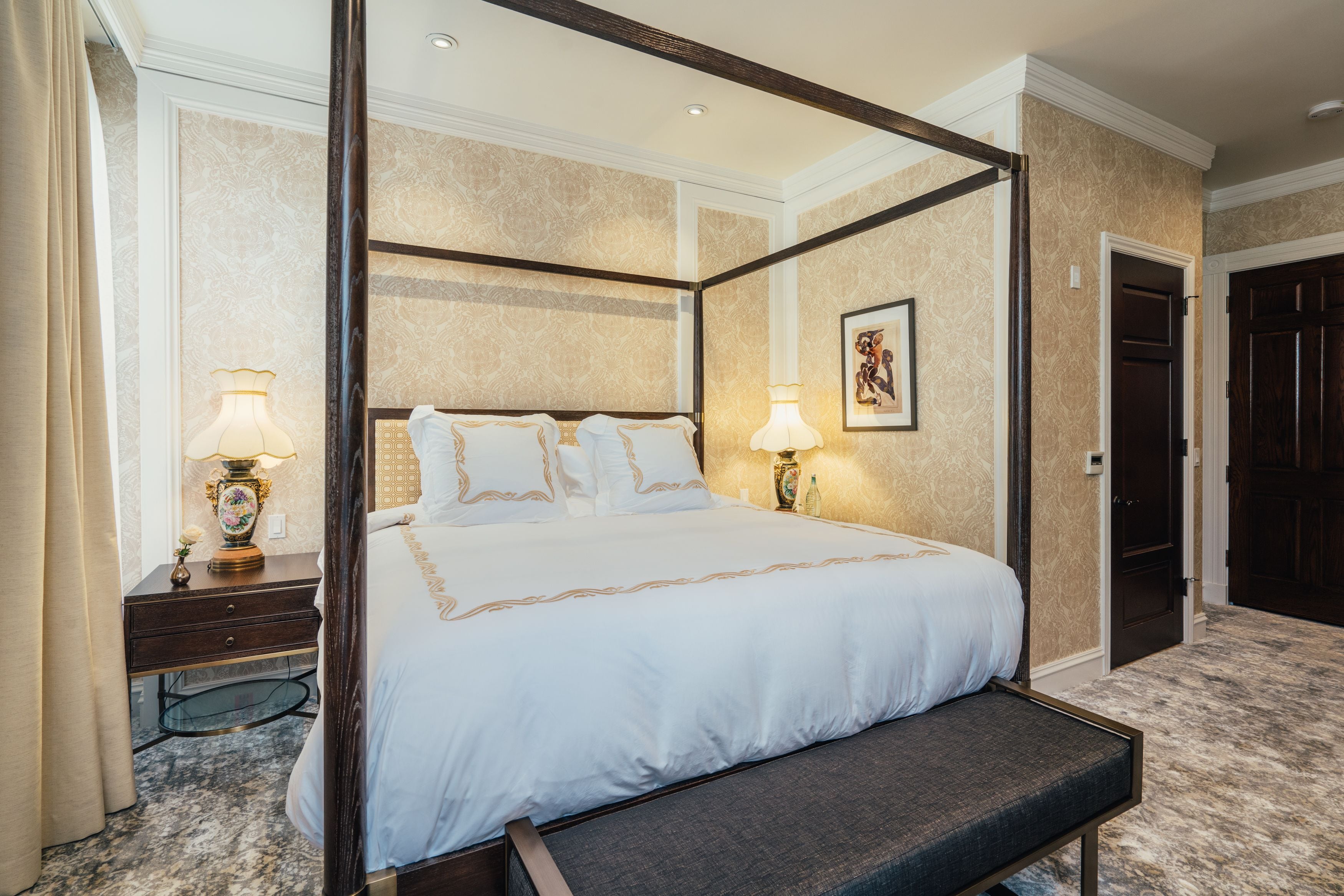 A four-poster bed with a bench, bedside lamps, and a framed artwork on the wall.
