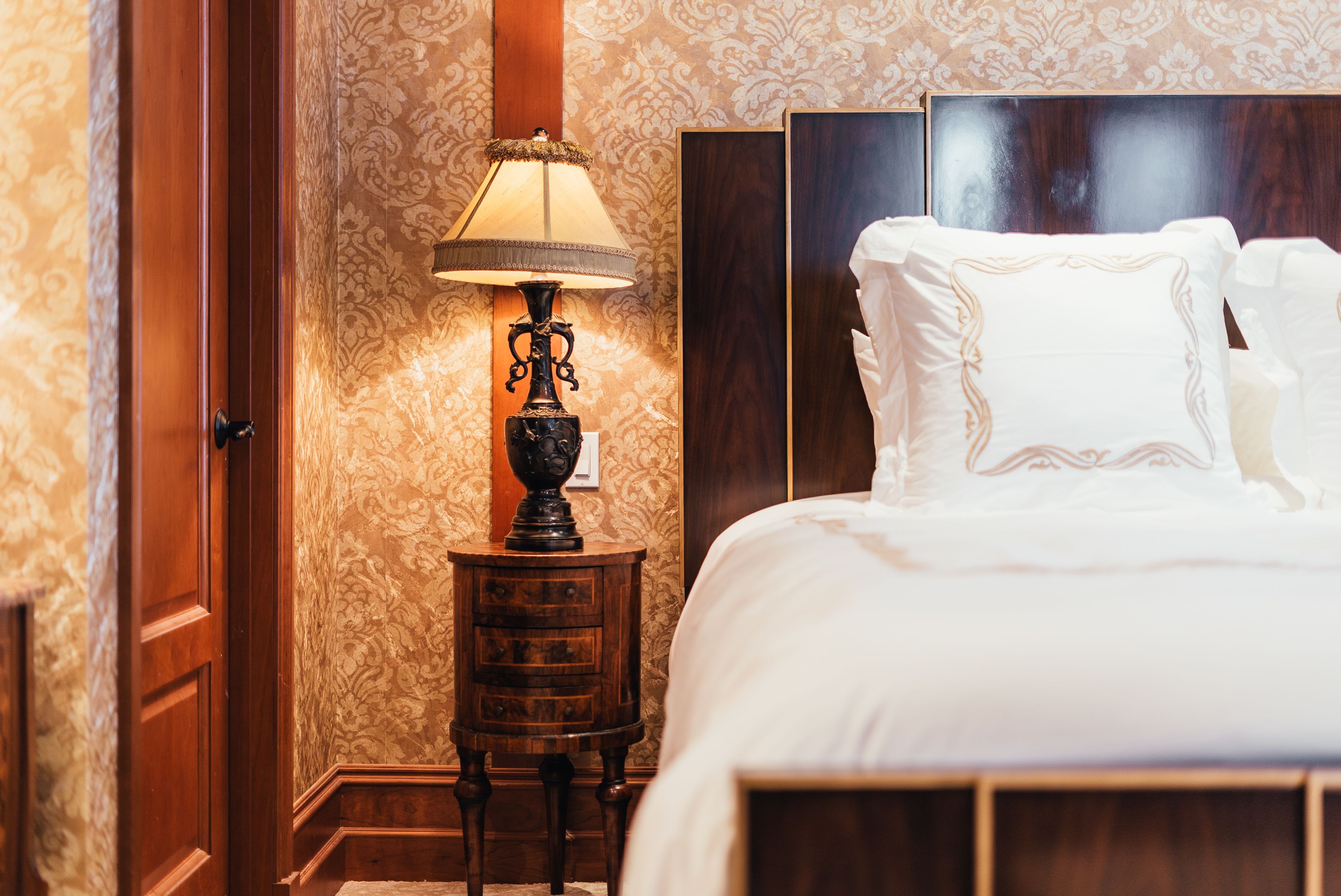 A neatly made bed with white linens and a wooden headboard, a small wooden nightstand with a lamp, and a door with a wooden frame.