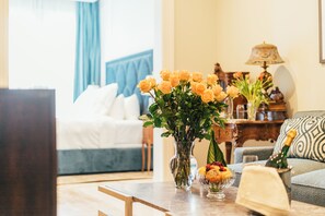 A hotel room with a bed, a sitting area with a sofa, a table with a vase of flowers, a fruit bowl, and a bottle of champagne. - Sutter Mansion (San Francisco)