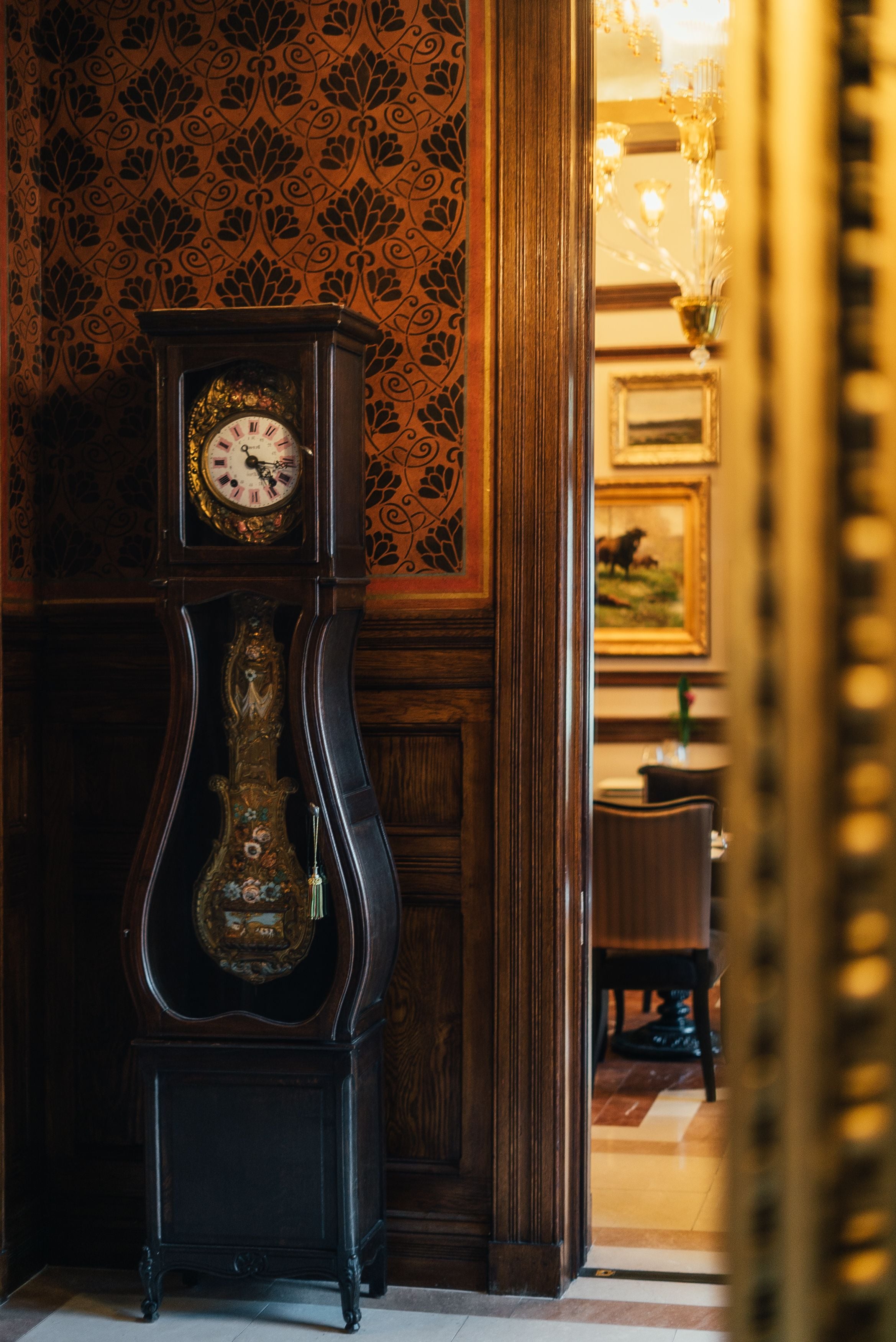 A grand, ornate grandfather clock with a detailed face and a decorative base, set against a richly patterned wallpaper and wooden paneling.