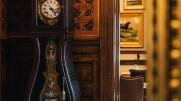 A grand, ornate grandfather clock with a detailed face and a decorative base, set against a richly patterned wallpaper and wooden paneling.