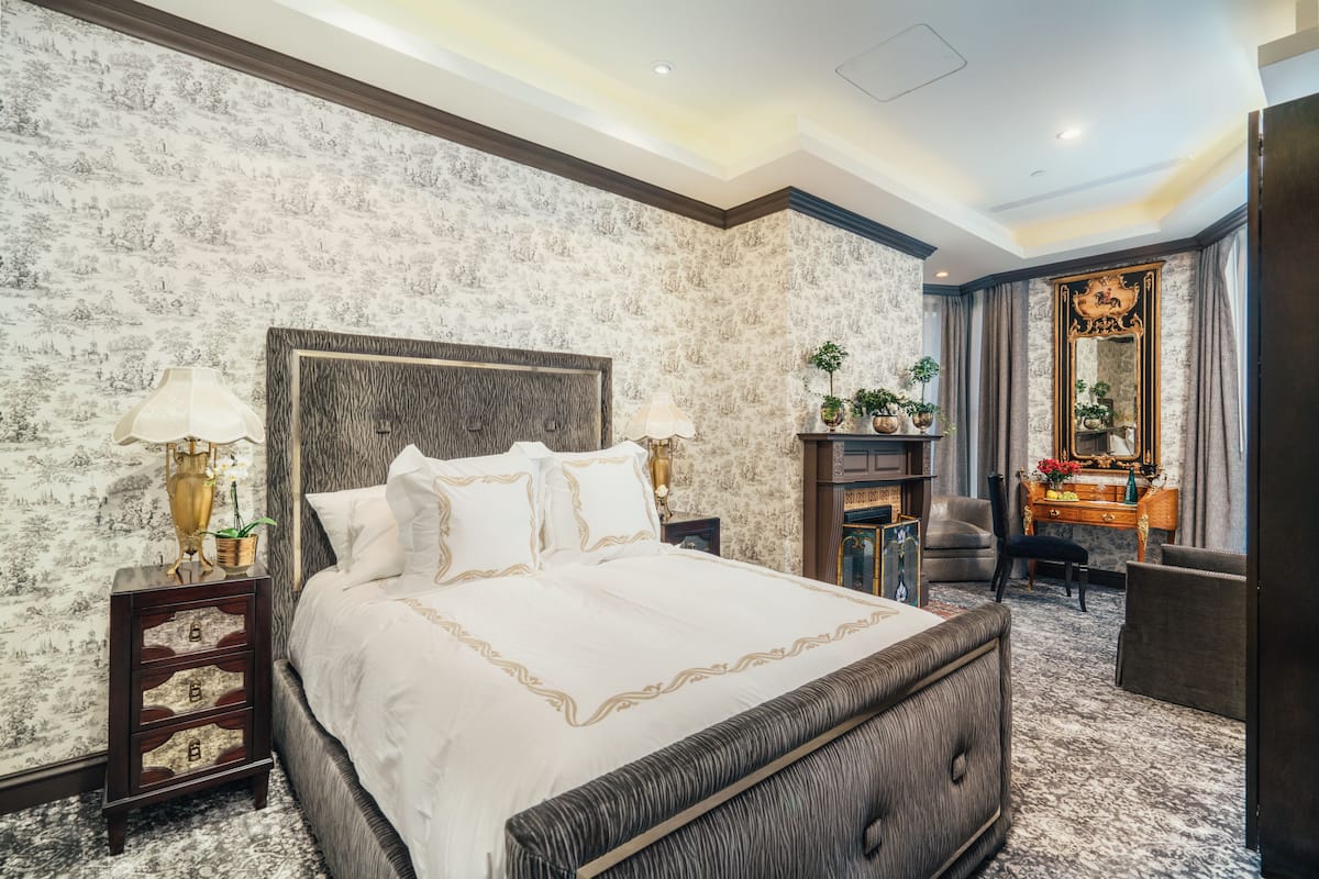 A spacious bedroom with a large bed, a nightstand with a lamp, a dresser, a mirror, and a seating area with a chair and a small table.