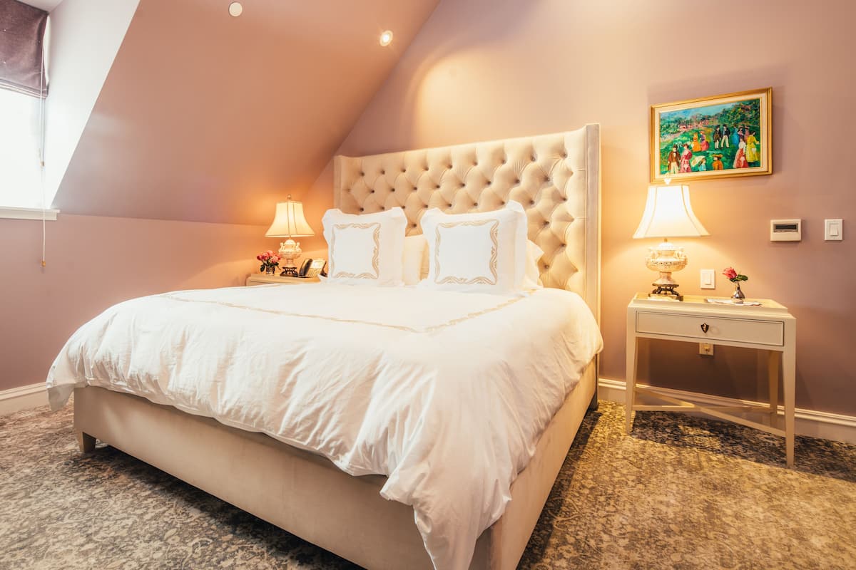A bedroom with a large bed, a tufted headboard, two bedside tables with lamps, a painting on the wall, and a carpeted floor.