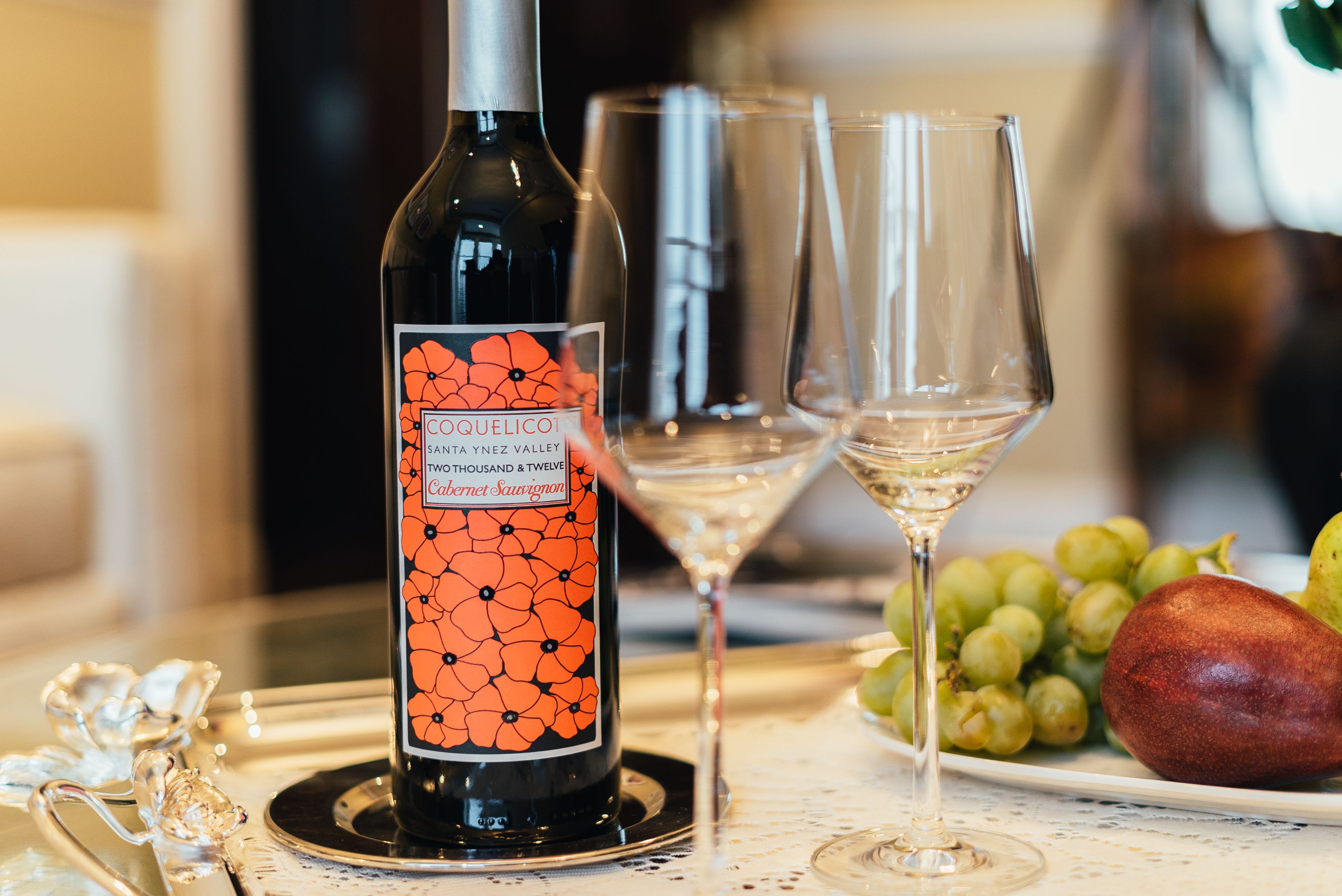 A bottle of Coquelico wine, two empty wine glasses, and a plate with grapes and a pear on a table.