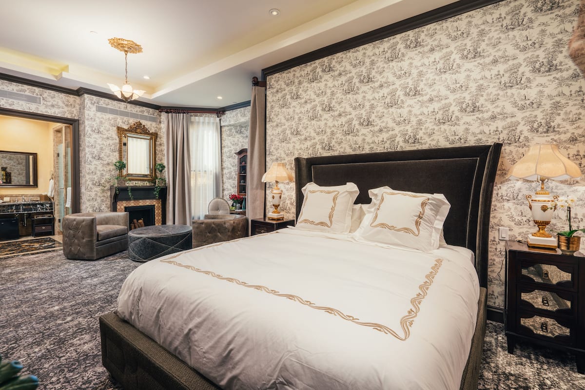A spacious bedroom with a large bed, a fireplace, a sitting area with a chair, and a vanity with a mirror.