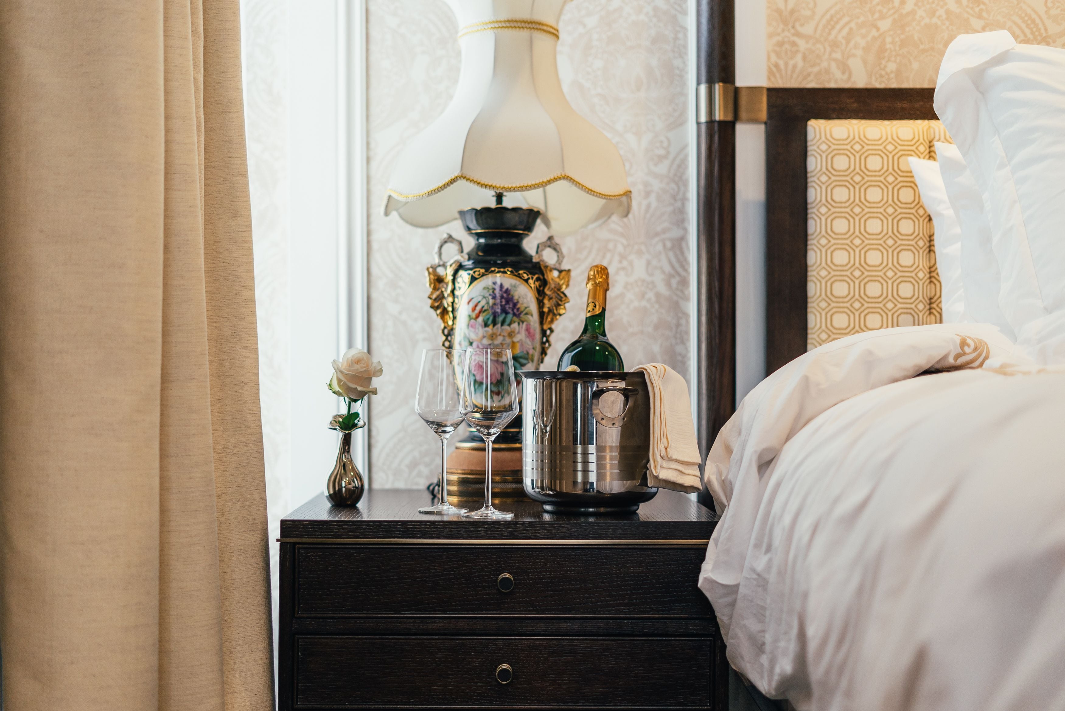 Una cama bien hecha con edredón blanco, una mesita de noche con lámpara, una botella de champán y un pequeño florero con una sola flor.