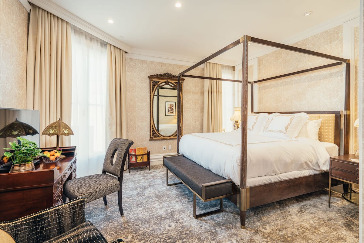 A four-poster bed with a bench, a desk with a lamp, a chair, and a mirror in a hotel room.