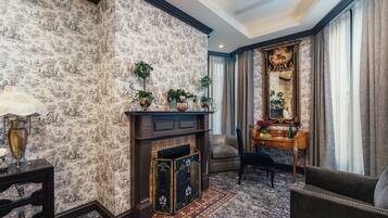 Quarto com papel de parede estampado, lareira com azulejos decorativos, mesa de cabeceira de madeira com abajur, uma cadeira e um espelho grande e ornamentado.