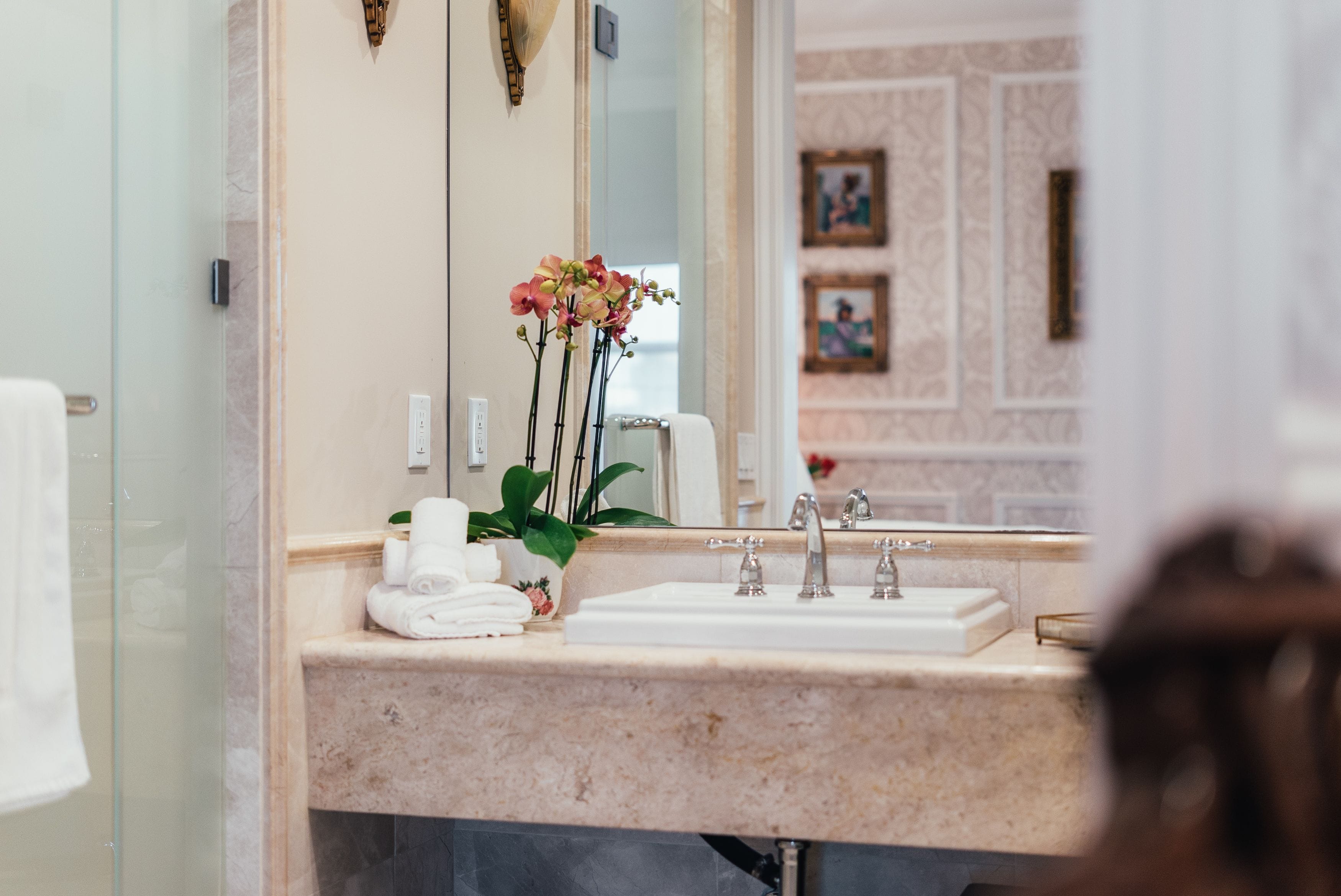 A bathroom with a marble sink, two faucets, a mirror, and a vase with flowers.