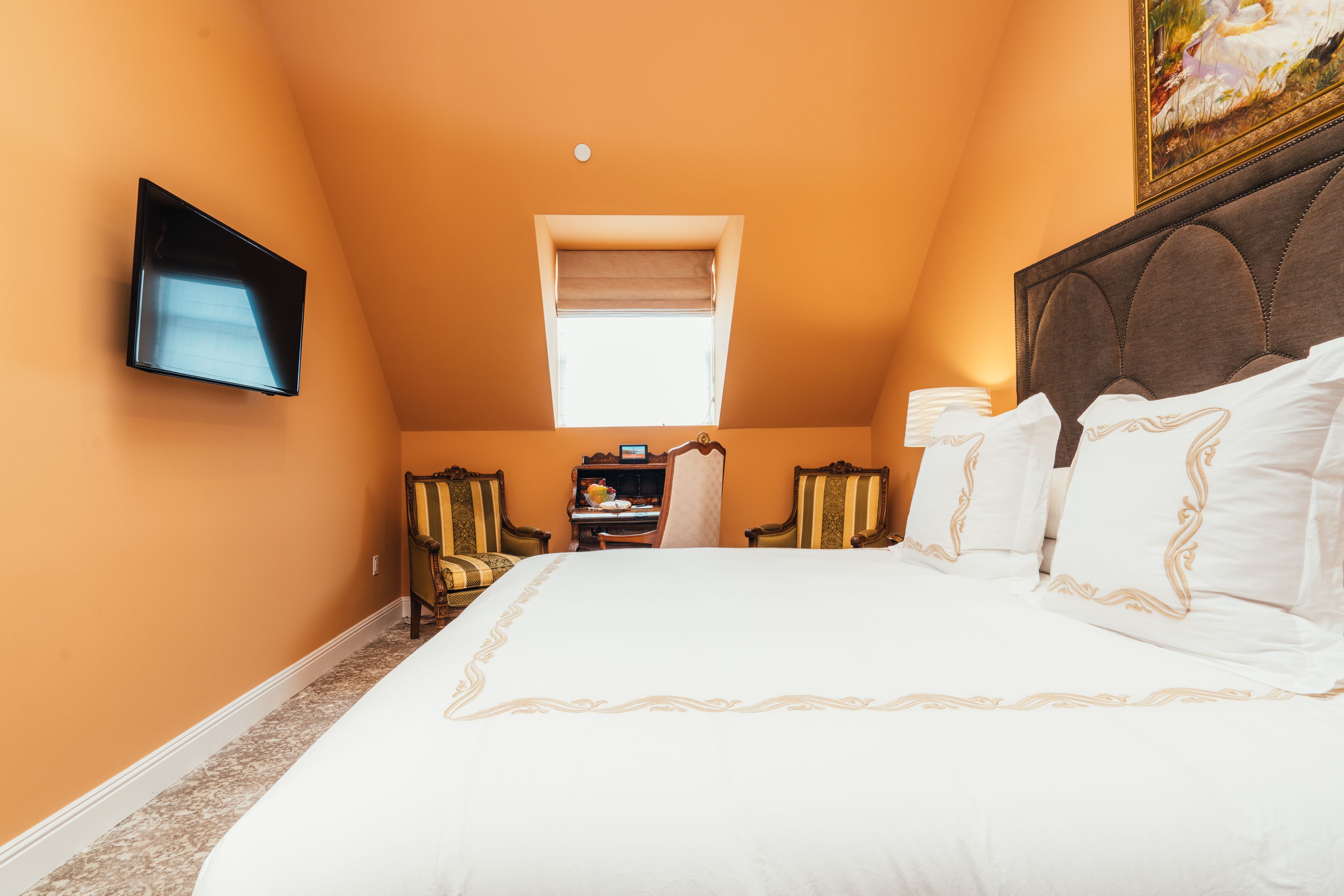 A bedroom with a large bed, a television mounted on the wall, a window with a blind, and a painting above the bed.