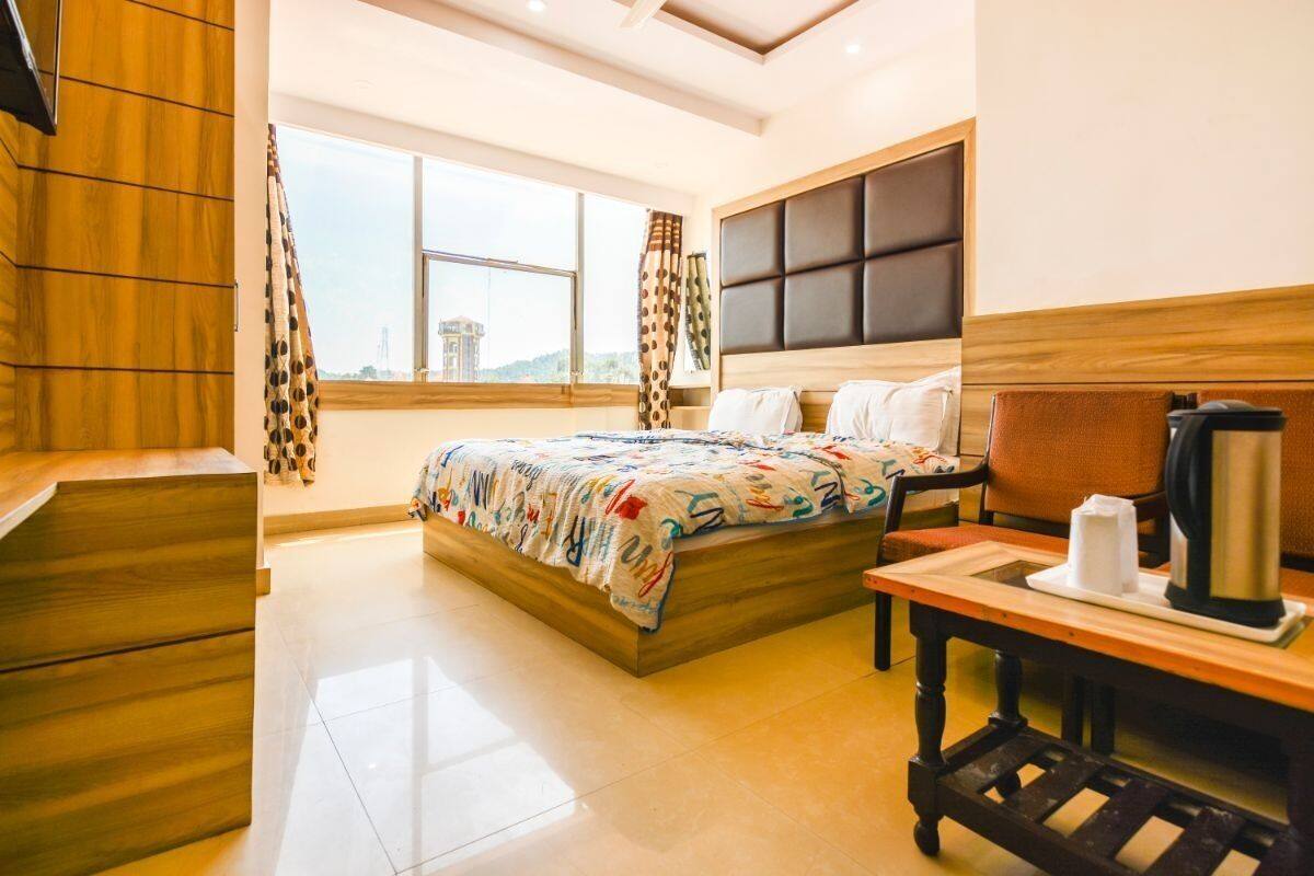 A hotel room with a large bed, a wooden headboard, a bedside table with a kettle, and a view of a cityscape through large windows.
