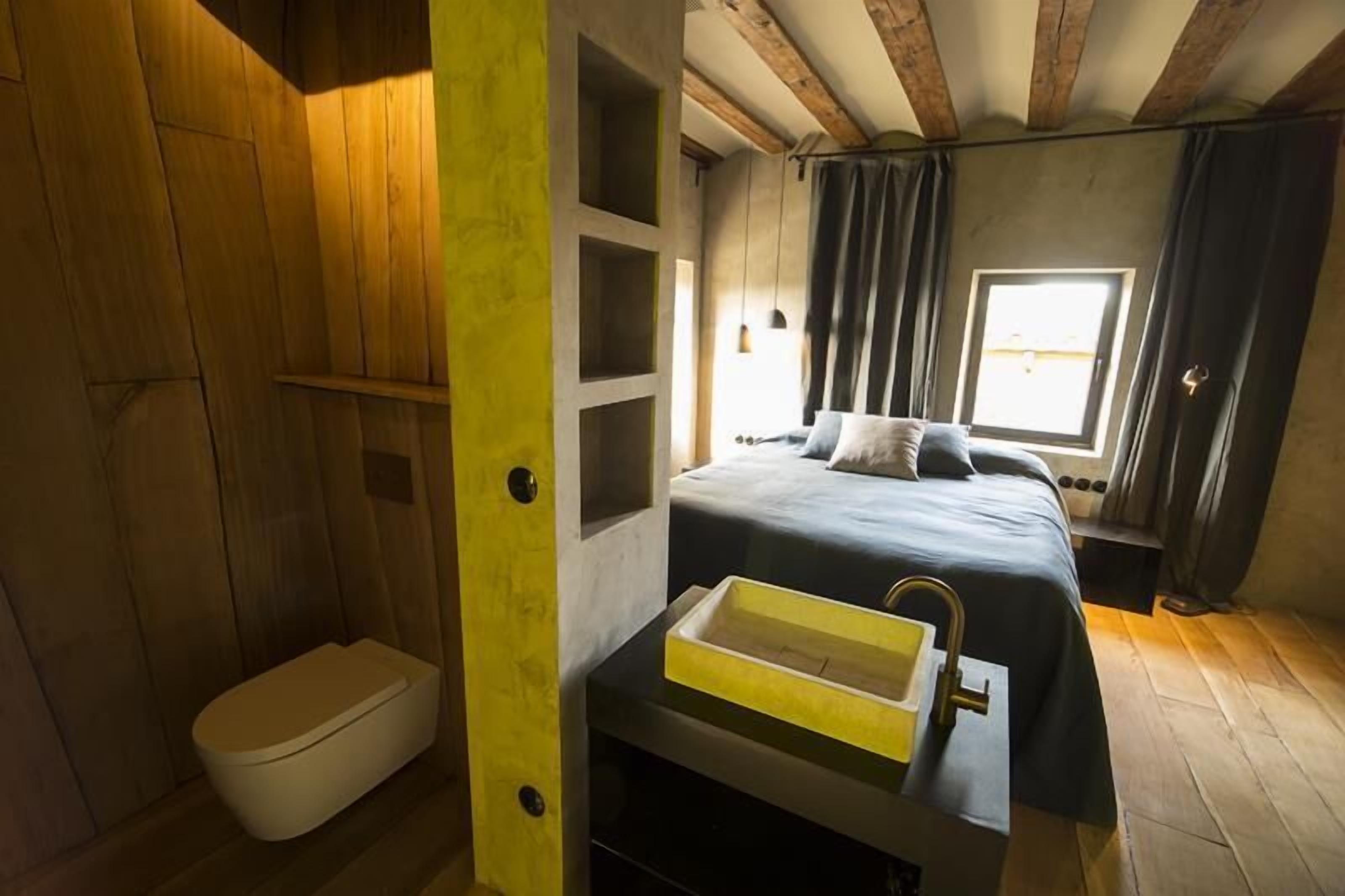 Un dormitorio con cama, ventana, estante empotrado y un baño pequeño con inodoro.