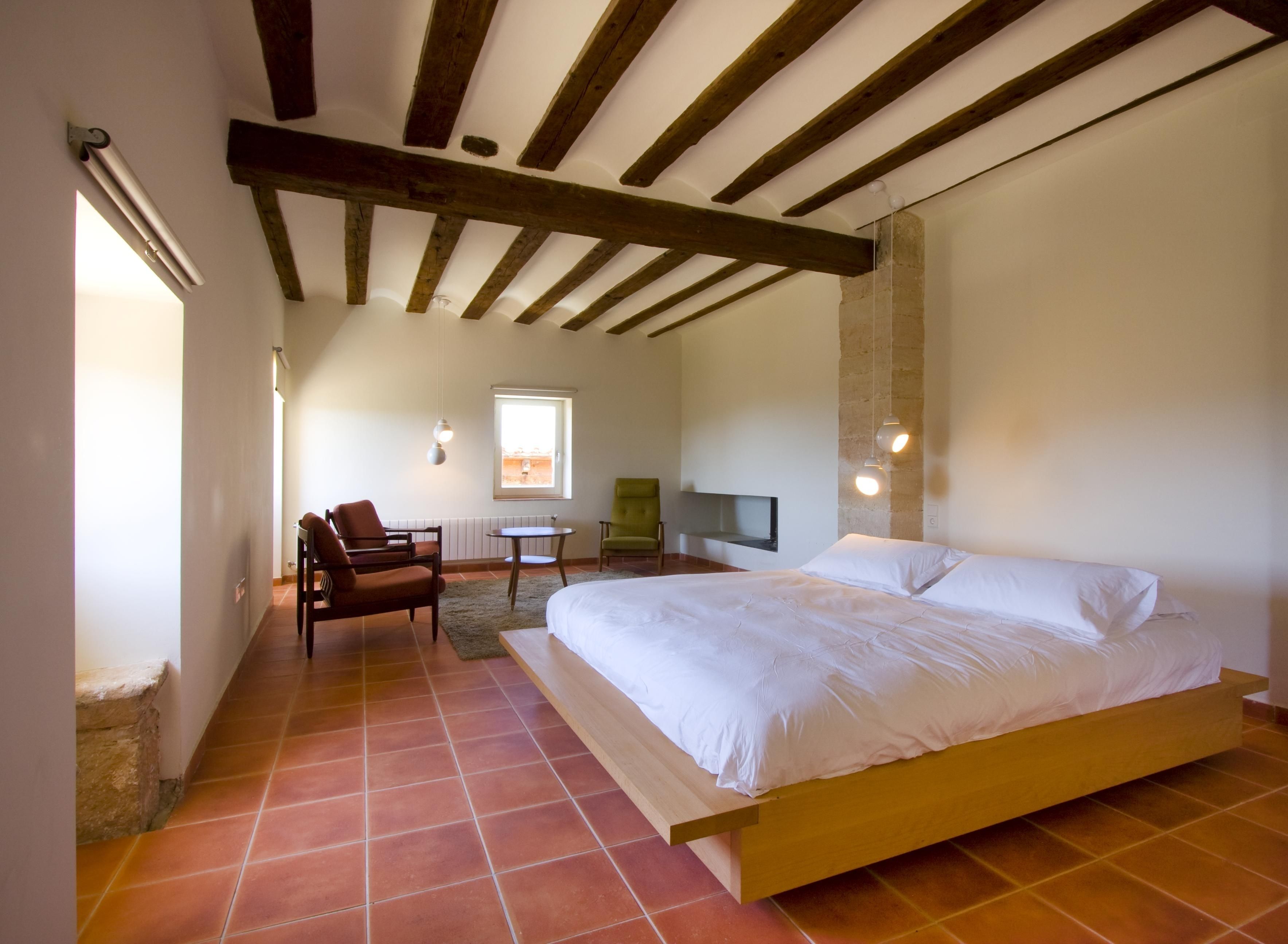 Un dormitorio con una cama de madera, una silla verde, una mesita pequeña y un televisor montado en la pared.