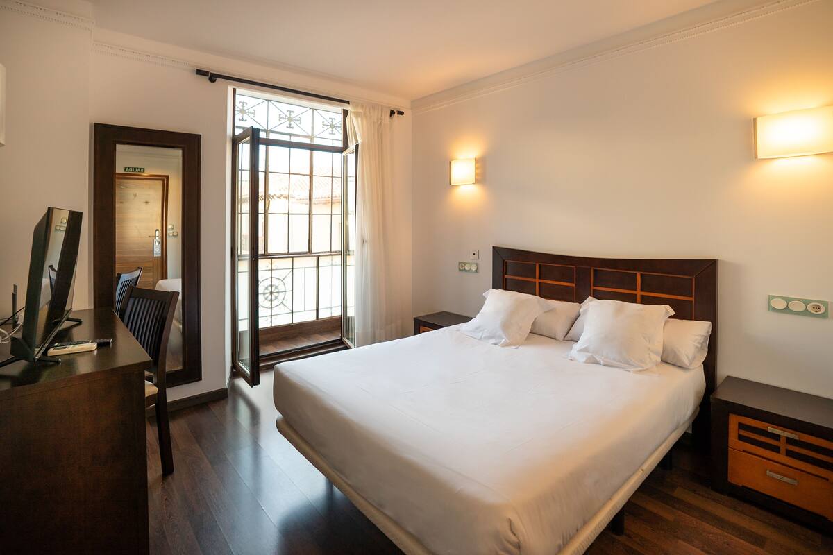 Una habitación de hotel con una cama grande, un escritorio, una silla, una mesita de noche y una ventana con cortinas.