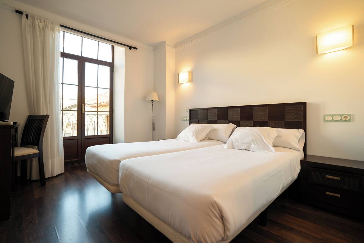 Habitación de hotel con dos camas, un escritorio, una silla, una lámpara y una ventana con cortinas.