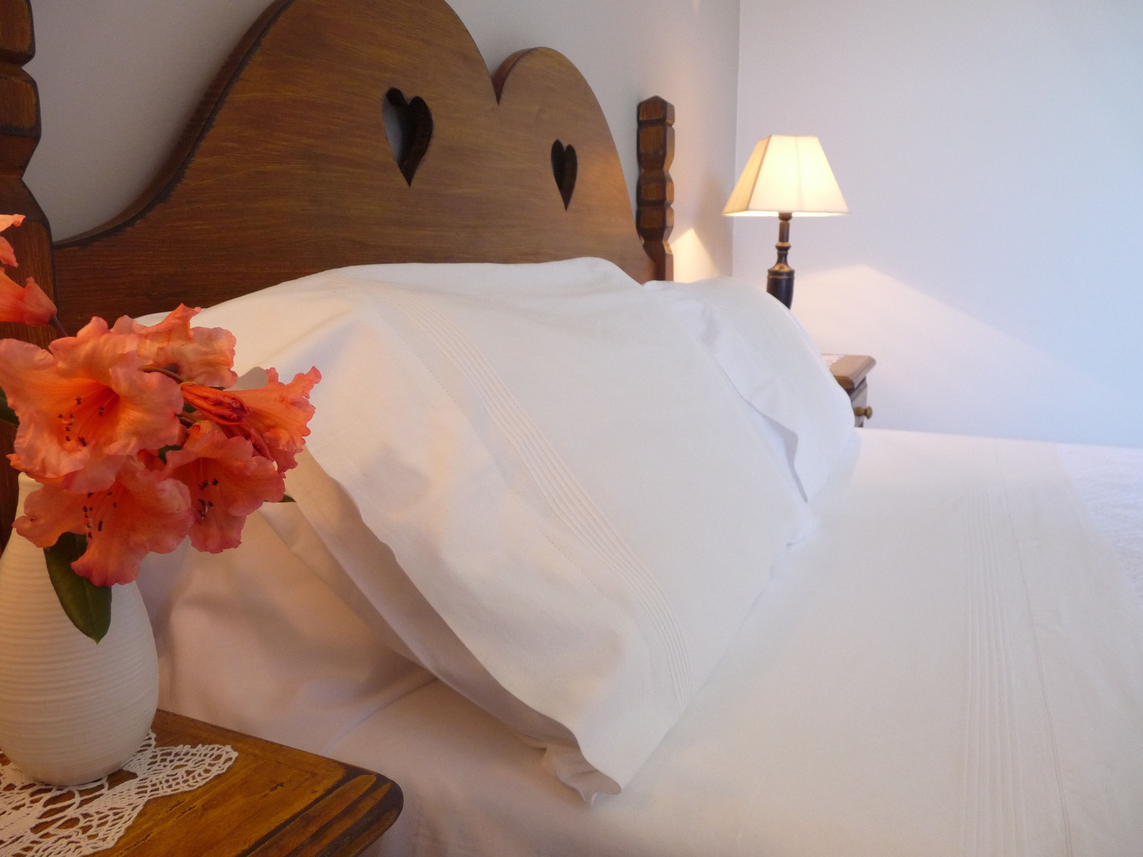 Een net opgemaakt bed met witte lakens, een houten hoofdbord met hartjesuitsparingen, een nachtkastje met een bloemenvaas en een bedlampje.