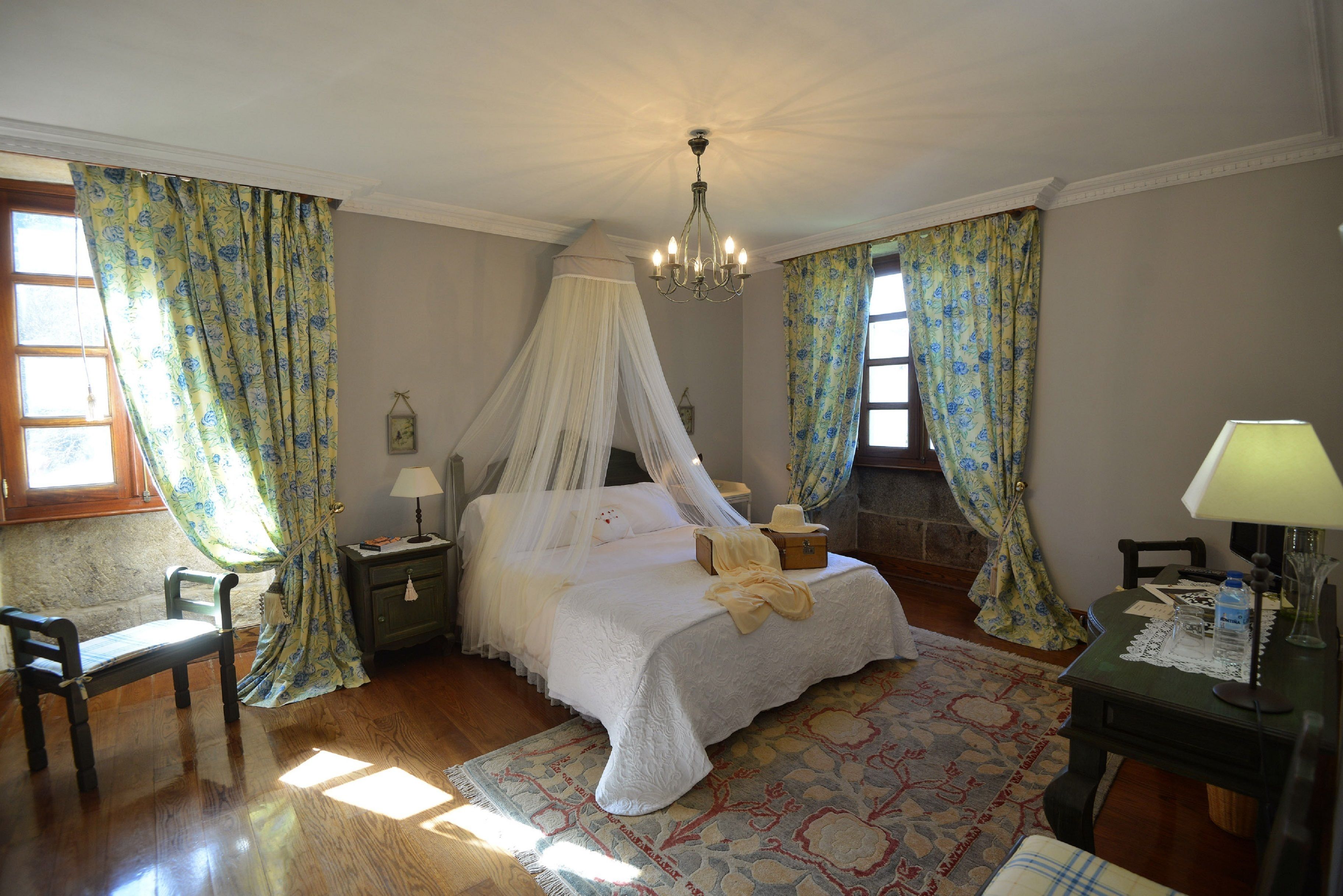 A bedroom with a four-poster bed, a nightstand, a chair, a table with a lamp, and a patterned rug.