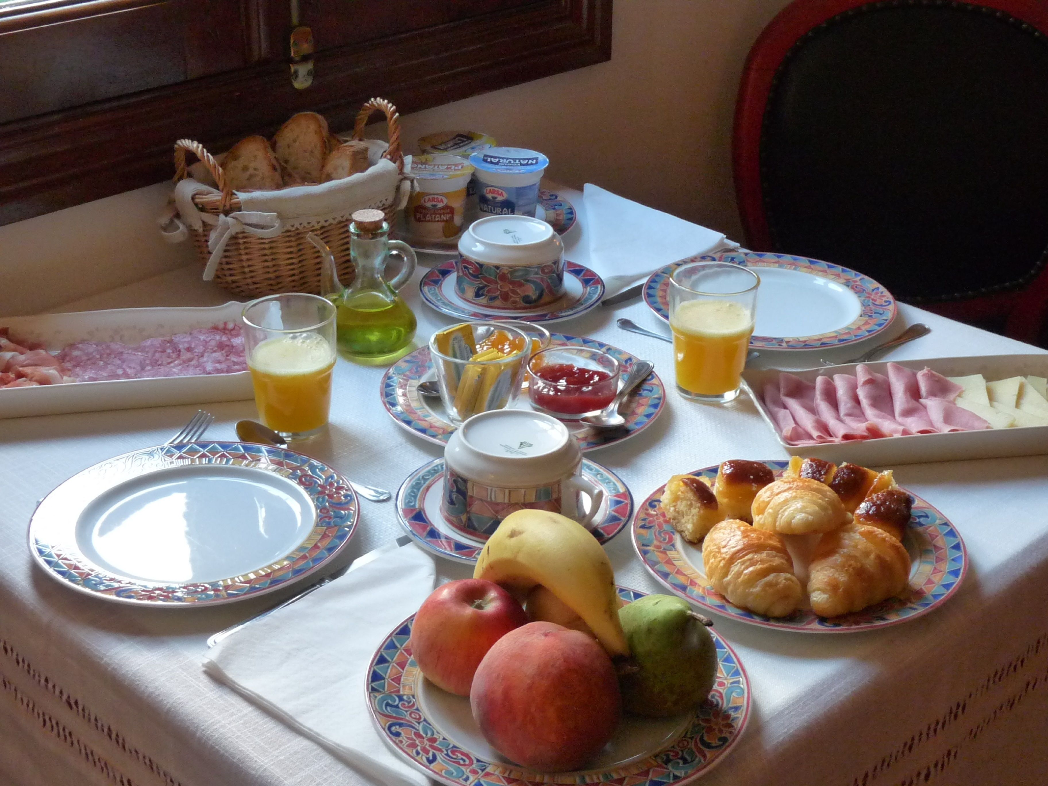 A breakfast table with plates of fruits, croissants, ham, and juice.