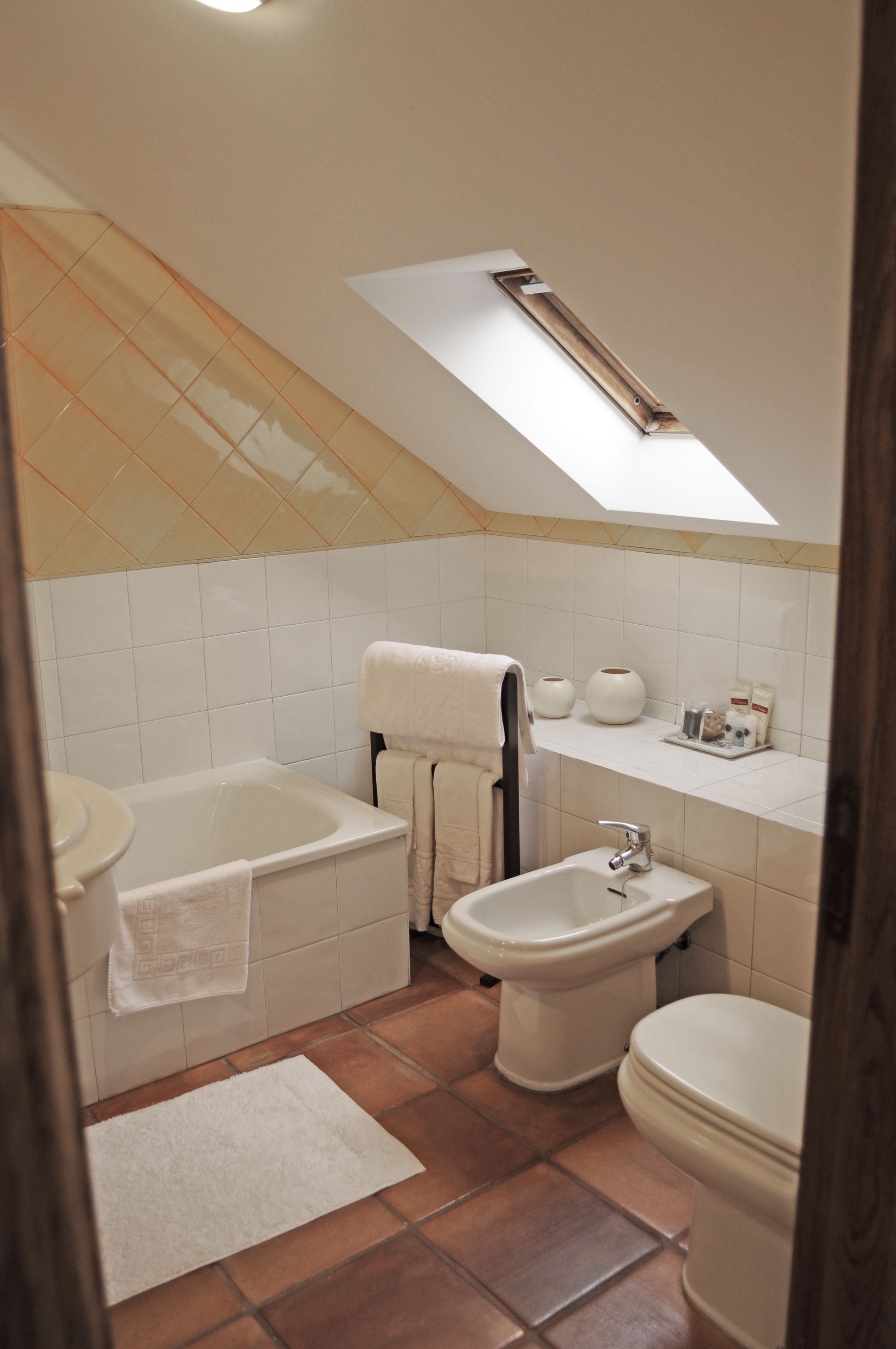 Une salle de bain équipée d’une baignoire, d’un lavabo, d’un bidet et de toilettes. Une lucarne se trouve au-dessus de la baignoire, et une étagère contient des articles de toilette.