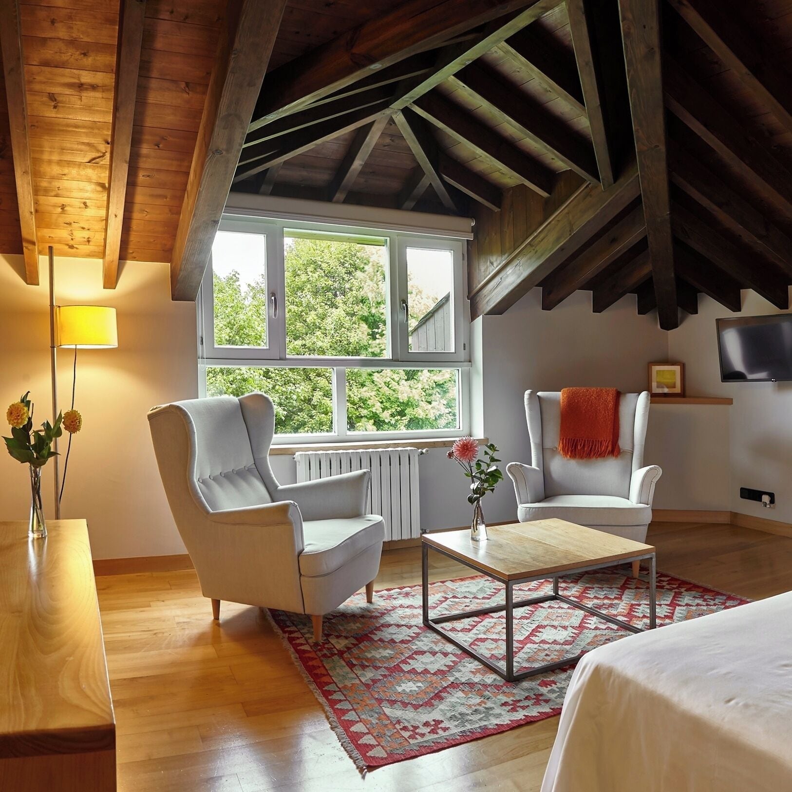 A room with a sloped ceiling, wooden beams, a large window, two white armchairs, a small table, and a bed.