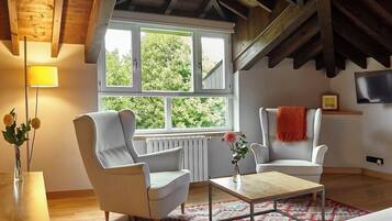 A room with a sloped ceiling, wooden beams, a large window, two white armchairs, a small table, and a bed.