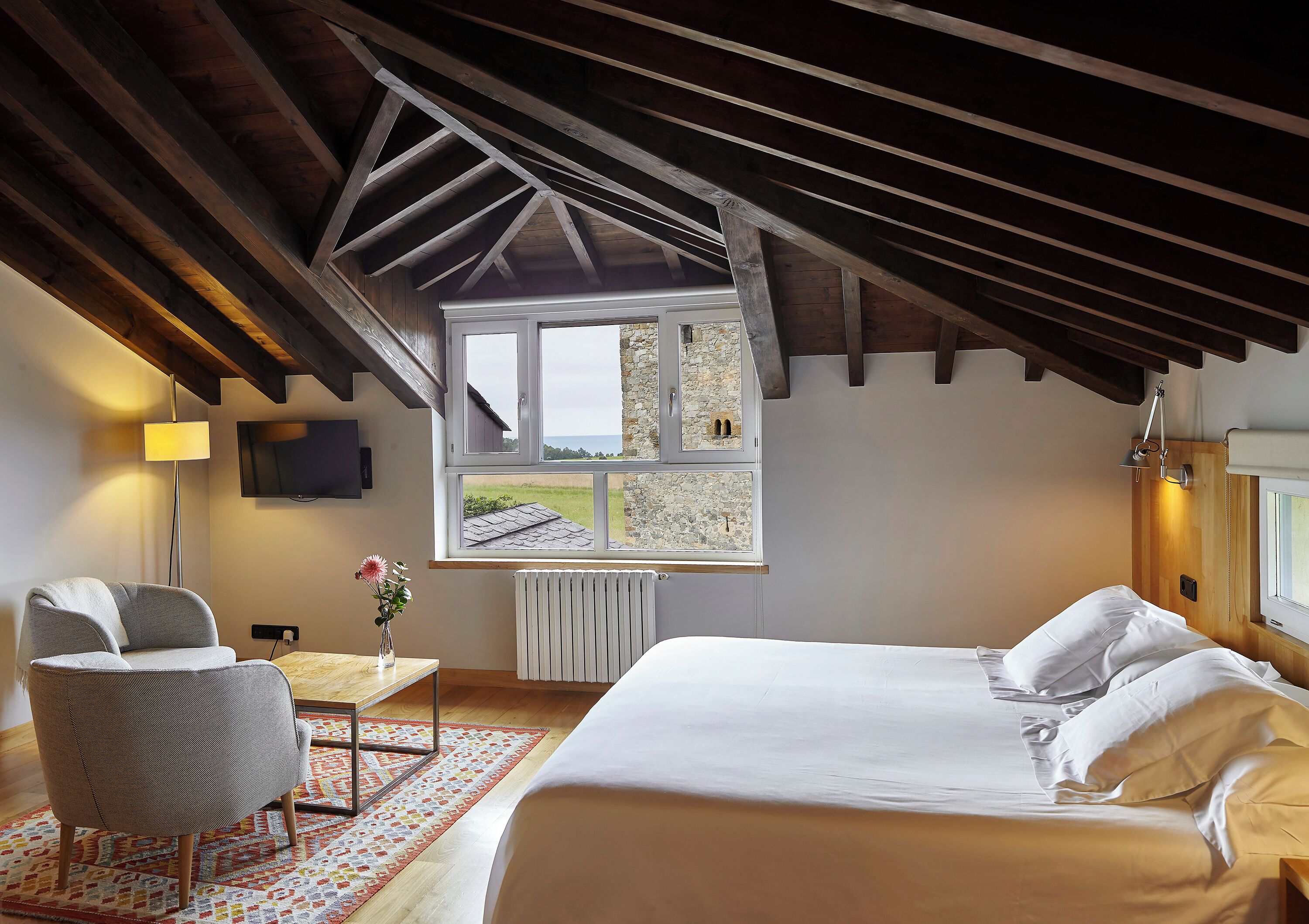 A bedroom with a large bed, a chair, a table, a TV, and a window with a view of the outside.