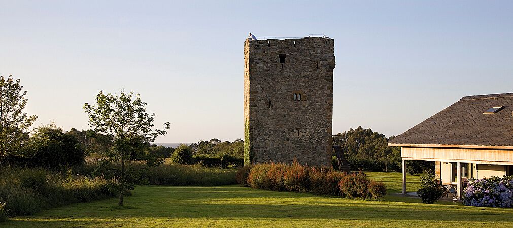 Una torre de piedra con techo plano, rodeada por un campo cubierto de hierba y un edificio con techo inclinado.