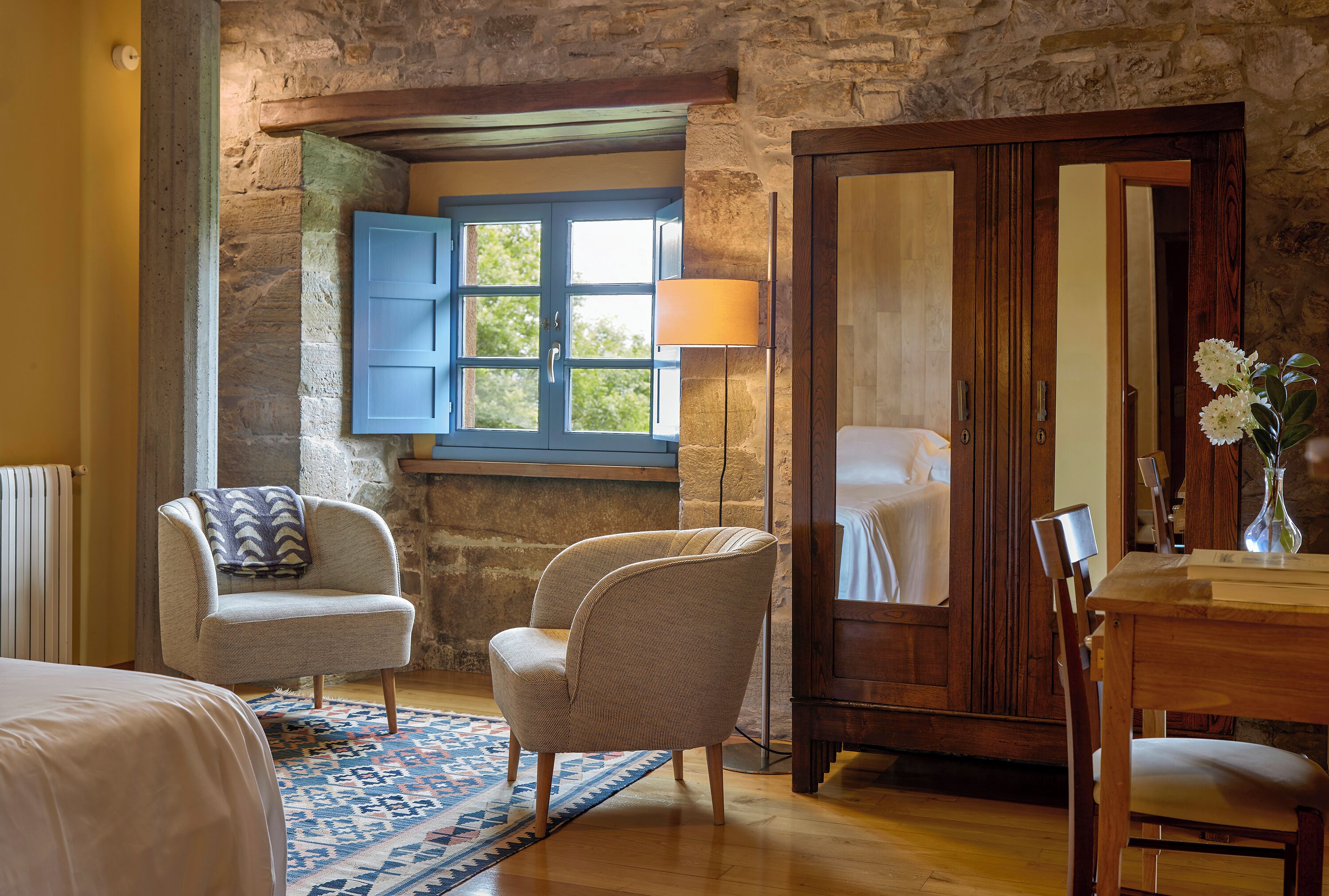 Una habitación con cama, dos sillas, un armario de madera, una ventana con contraventanas azules y una alfombra estampada.