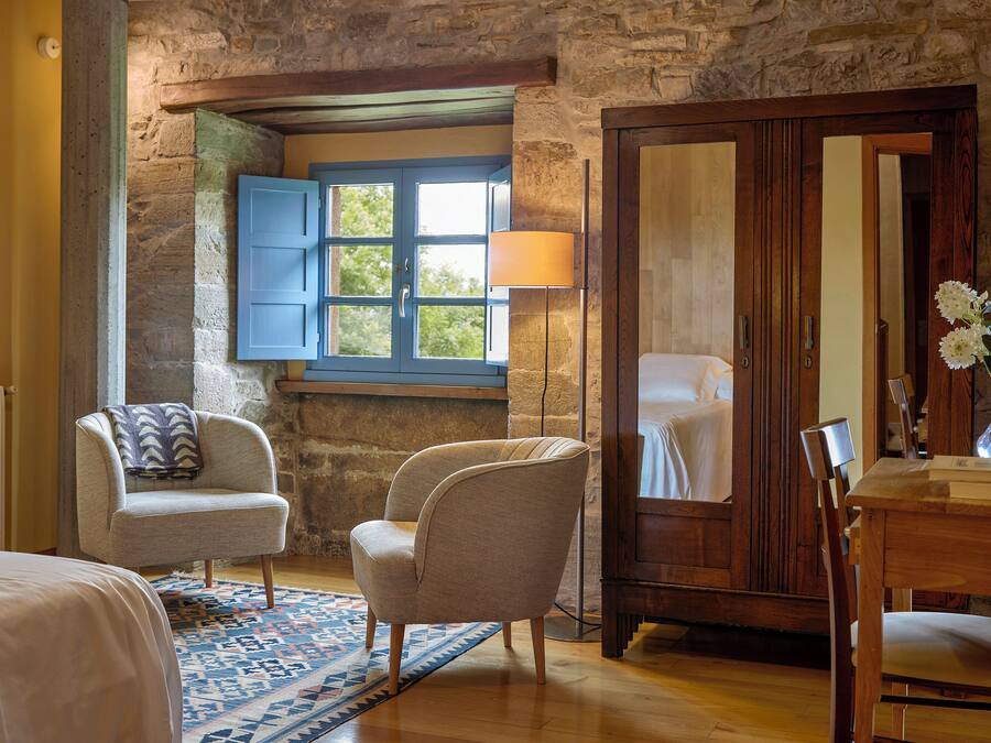Una habitación con cama, dos sillas, un armario de madera, una ventana con contraventanas azules y una alfombra estampada.