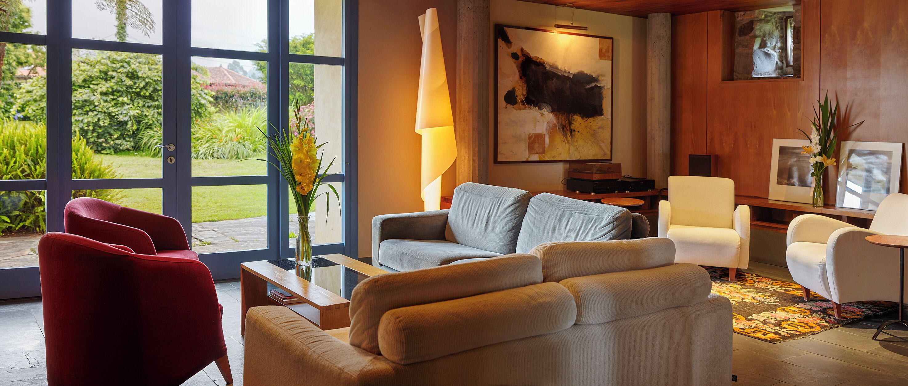A modern living room with a sofa, armchairs, a coffee table, and a lamp. There is a large window with a view of greenery outside.
