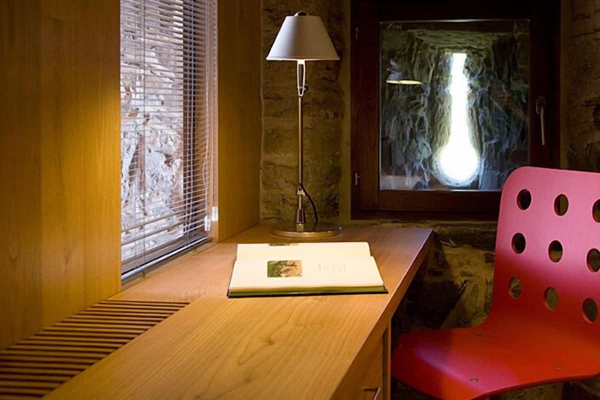Een kamer met een houten bureau, een rode stoel, een lamp, een raam met jaloezieën en een stenen muur.