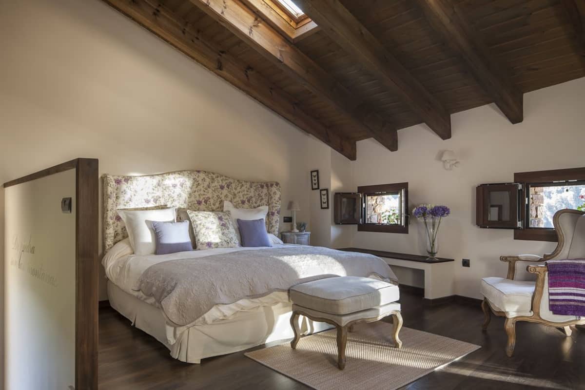 Une chambre avec un grand lit, des poutres en bois, une lucarne, une table de chevet avec une lampe, une chaise et un miroir.