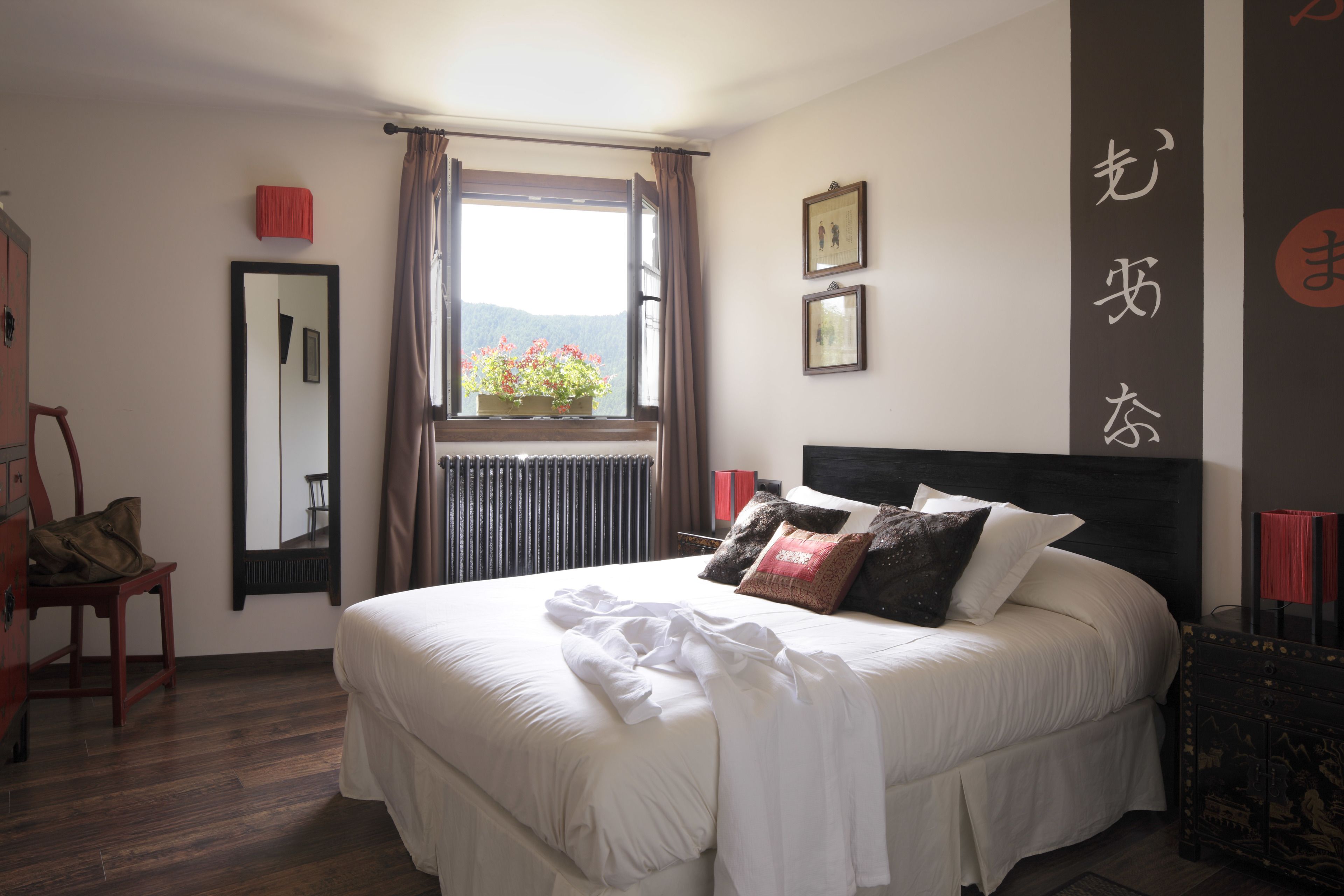 A neatly made bed with white linens, a window with a view of mountains, and a red chair.