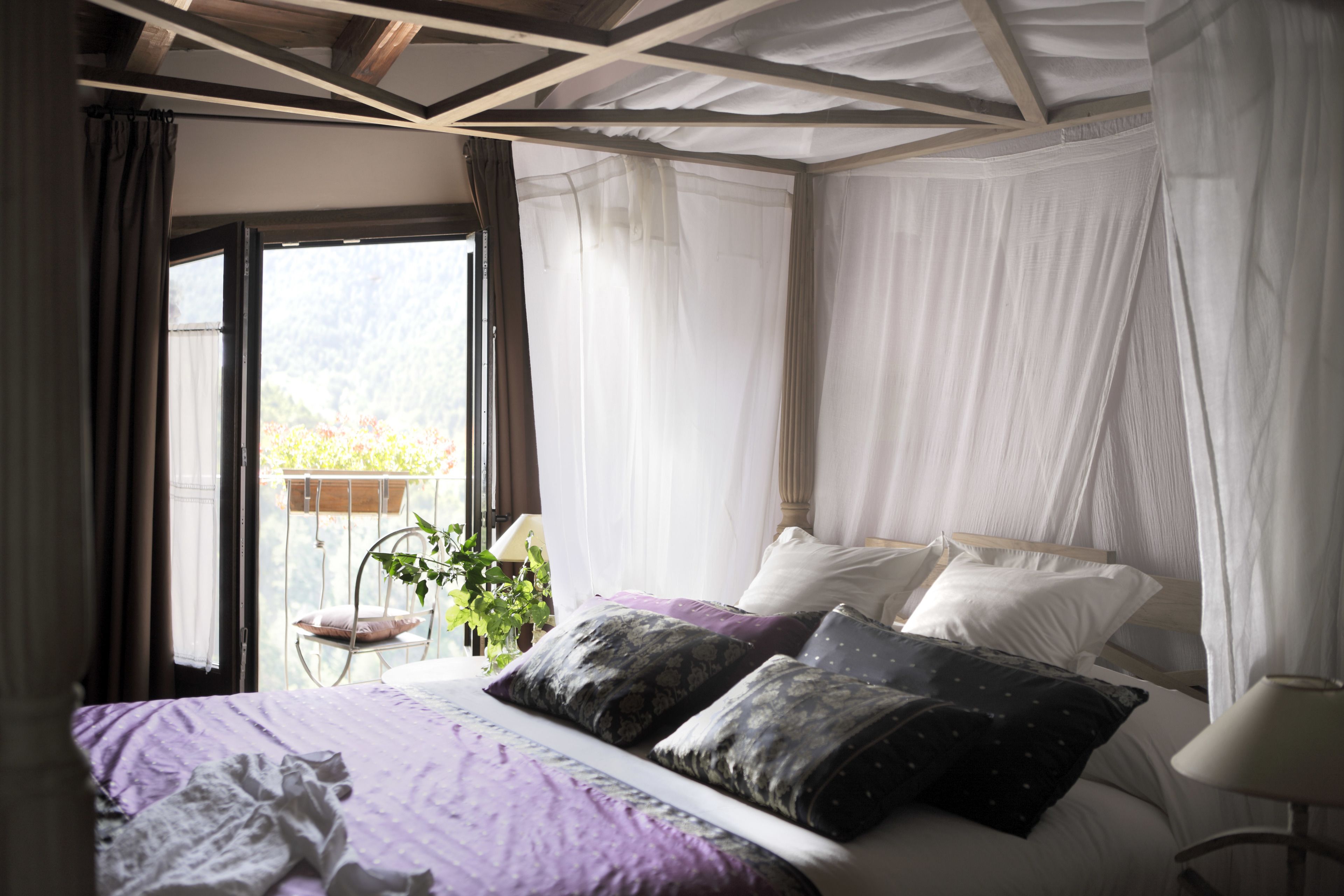 A bedroom with a bed, a window with curtains, a bedside table with a lamp, and a view of greenery outside.