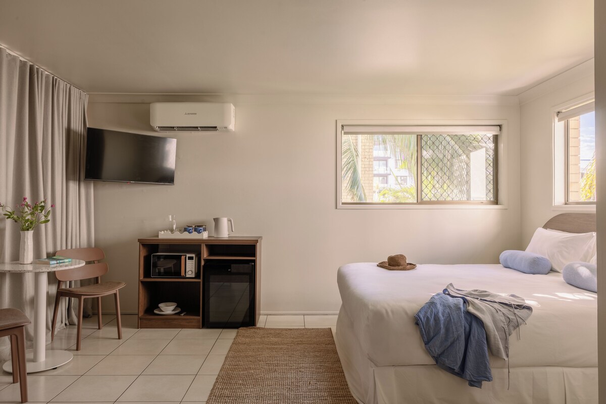 Een hotelkamer met een bed, een aan de muur bevestigde televisie, een klein tafeltje met stoelen, een boekenplank en een raam met uitzicht op gebouwen.