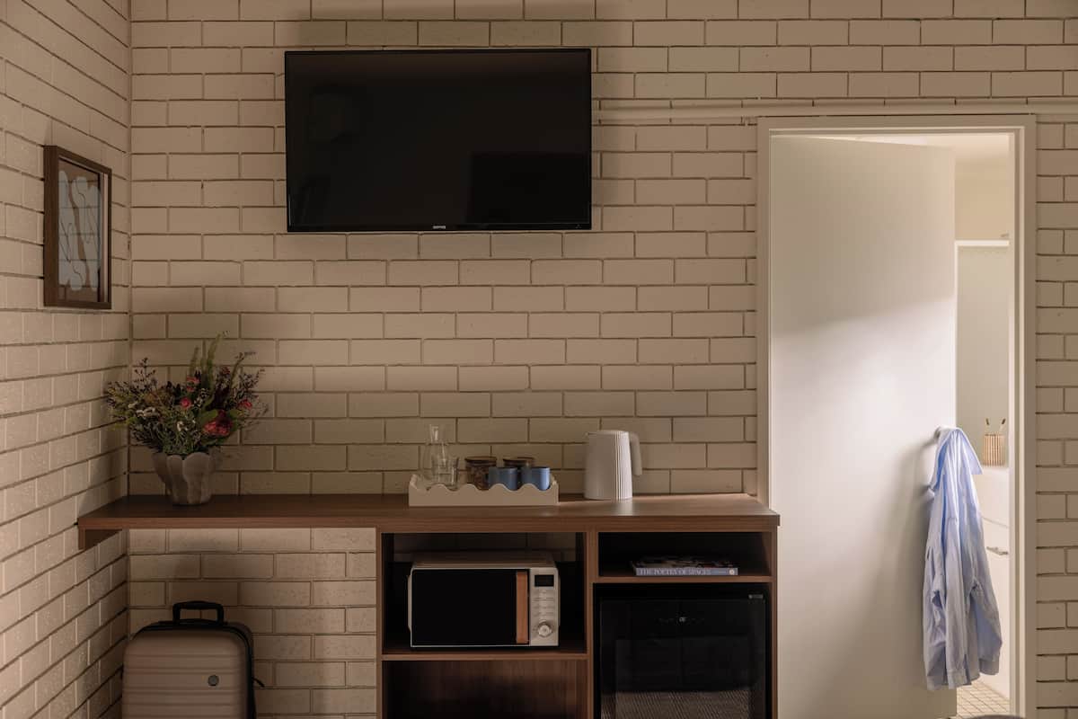 Une pièce avec un mur en briques, un téléviseur à écran plat, une étagère en bois ornée d’un vase de fleurs, un four à micro-ondes, une valise et une porte sur laquelle est accrochée une serviette.